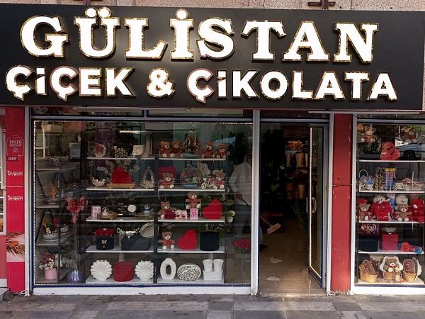 Gülistan Çiçek & Çikolata dükkânının cepheden görünüşü; siyah tabelada beyaz ışıklı harfler, vitrinde oyuncak ayılar ve çikolata kutuları
